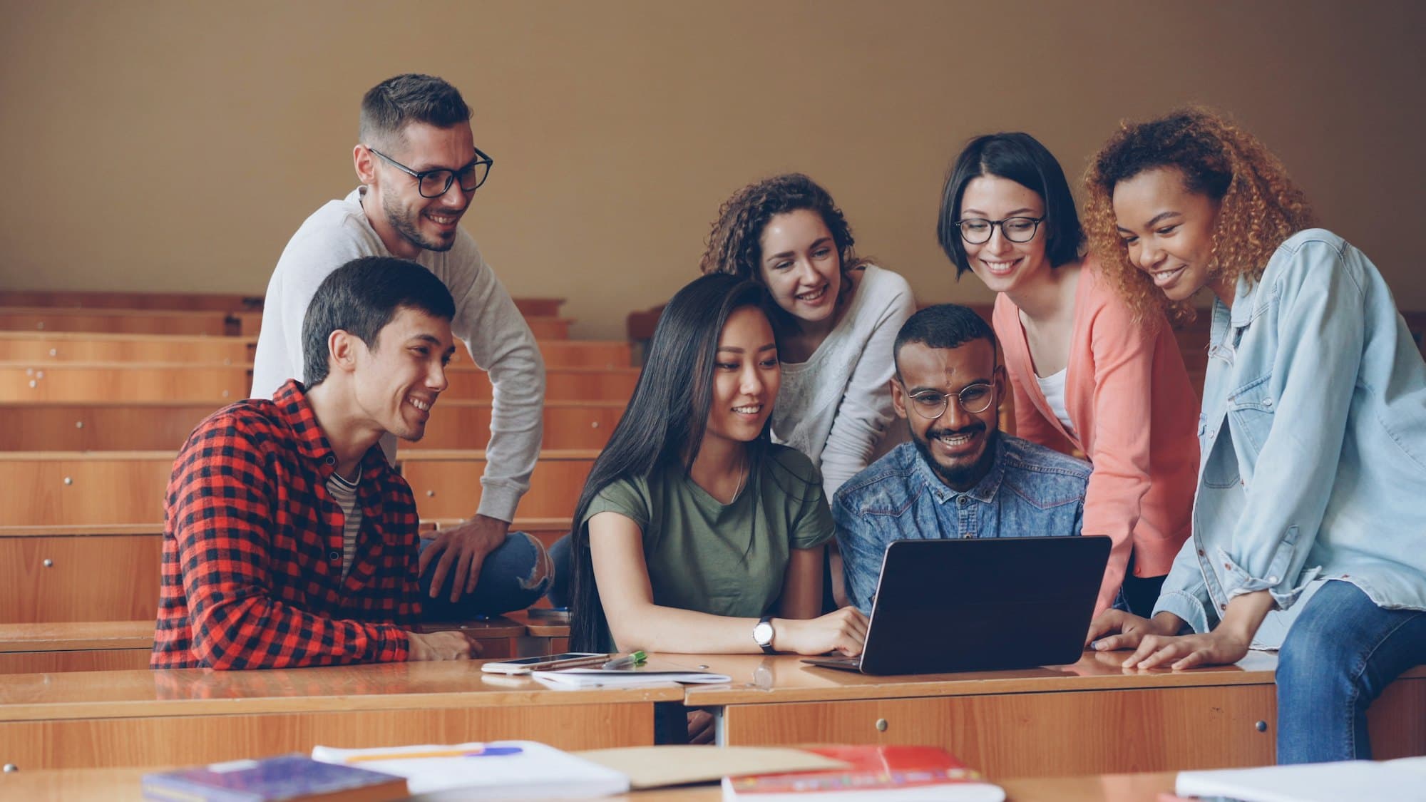 Students gathered around a laptop in a lecture hall, laughing